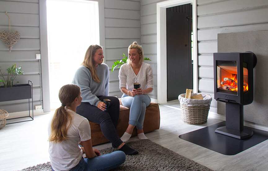 Three girls sitting on a brown pouf in a gray living room in front of the Jøtul F 167 wood stove